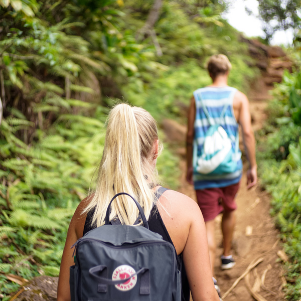 Two people wearing backpacks and tank tops walking on a forest trail 