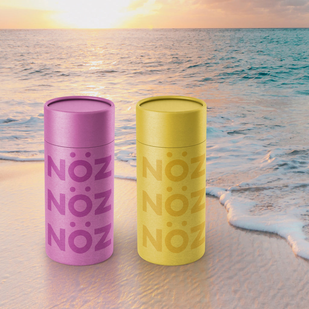 A purple and a yellow stick of Nöz sunscreen on the beach at sunset